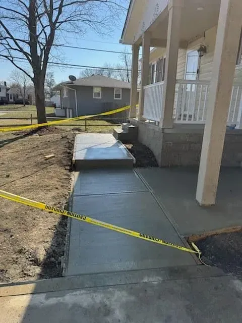 Newly poured concrete walkway leading to a house with yellow caution tape, overcast sky.