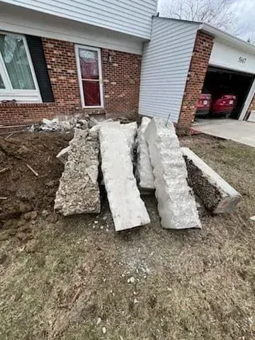 Concrete blocks piled on a grassy area near a house with a garage and a red car inside.
