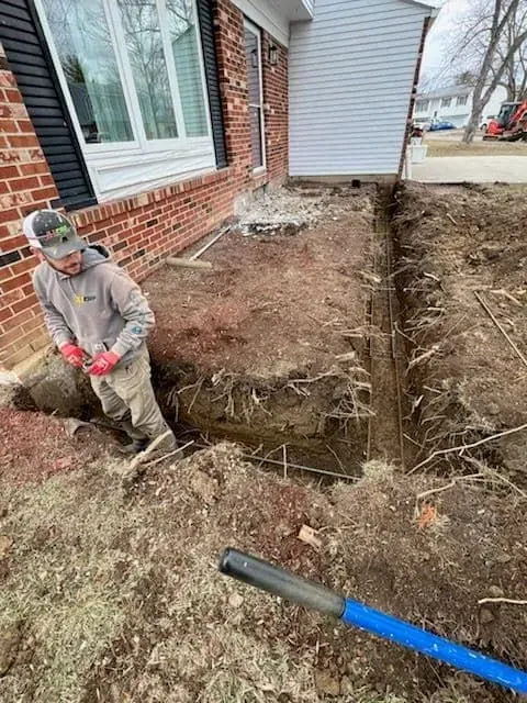 Person in a trench near a house with exposed foundation, preparing to lay a pipe.