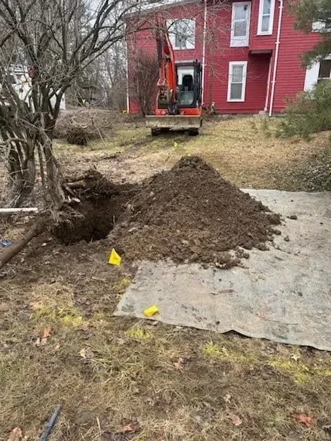 An excavator digs a hole in a yard near a red house. A pile of dirt and yellow flags are present.
