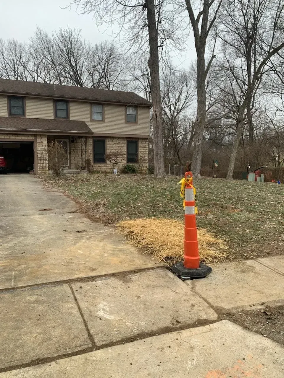 Orange traffic cone on sidewalk near a two-story house with bare trees.