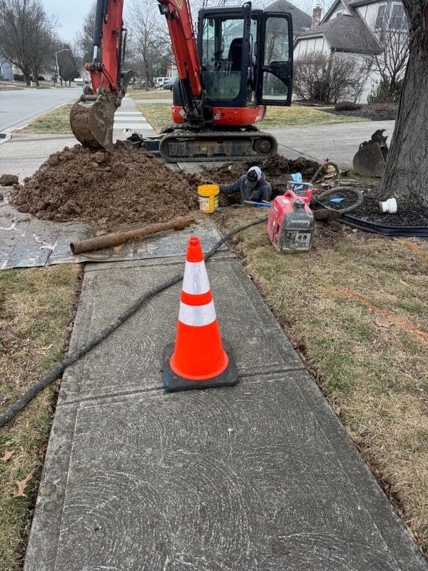 Excavator digging near a sidewalk; worker, orange cone, equipment, and dirt.