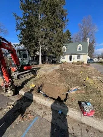 Excavator digs in front yard of house with green roof. Pile of dirt and construction equipment visible. Bright, sunny day.