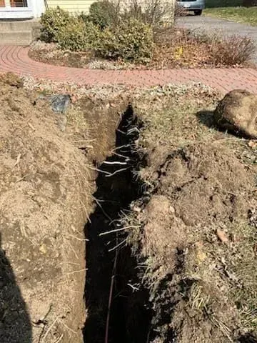 A deep trench dug in the ground in front of a house, exposing roots and a thin cable.