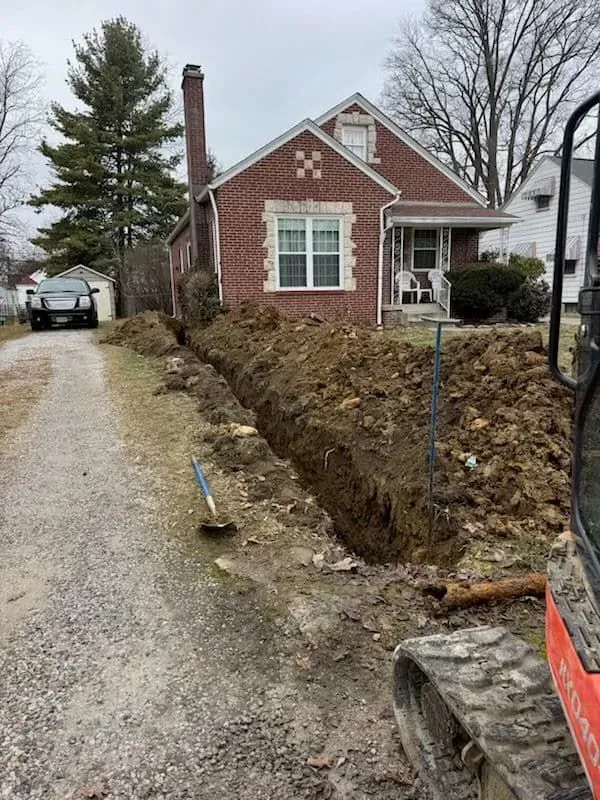 Trench dug next to a brick house; a small excavator is in the foreground, parked on the side of a driveway.