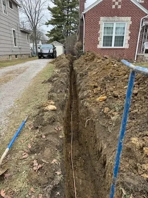 A trench dug alongside a driveway, likely for utilities. Brown dirt and a brick house are visible.
