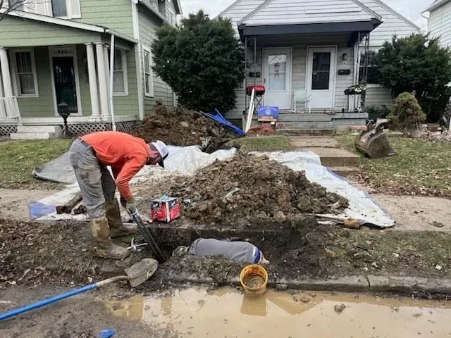 Two workers repairing a utility line in a residential street. One digs, the other works in the hole. Dirt pile visible.