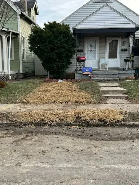 A house with a small front yard covered in dried grass, a tree and a few steps leading to a door.