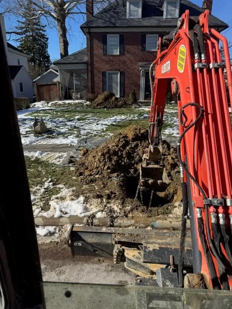 Red excavator digging in front yard of a brick house with patches of snow.