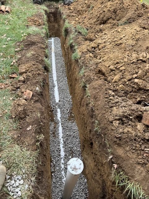 Trench dug in the ground with gray gravel and white pipe running through it.