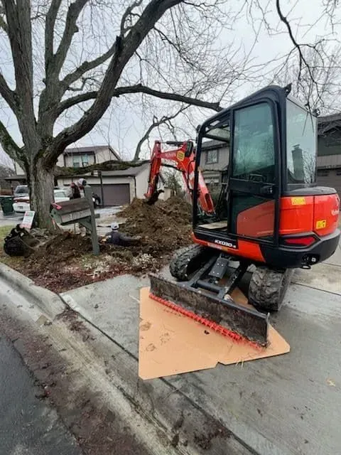 Orange excavator on a sidewalk, digging near a tree and a house.