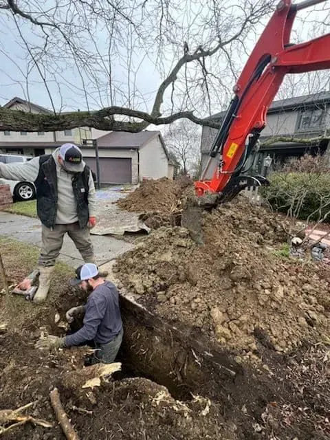 Two men working on a trench with an excavator in a residential area. One operates the machine, the other works in the trench.