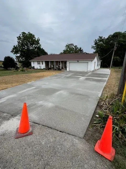 Newly poured concrete driveway in front of a white house, two orange cones in foreground, cloudy sky.