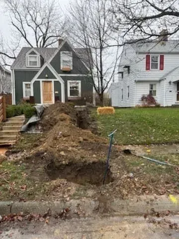 A house with a dirt excavation in front. Shovel and dirt pile in the yard; a partially wooden staircase is visible.