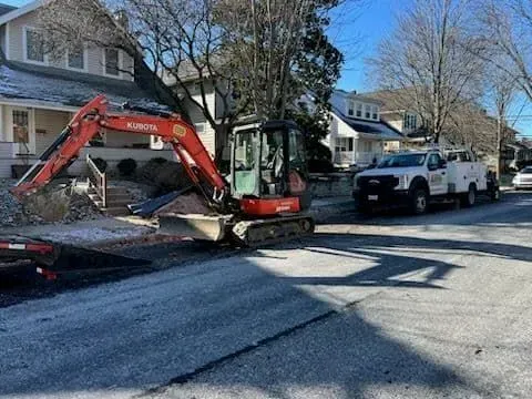 An orange excavator digs in a street next to a work truck, residential area with frost.