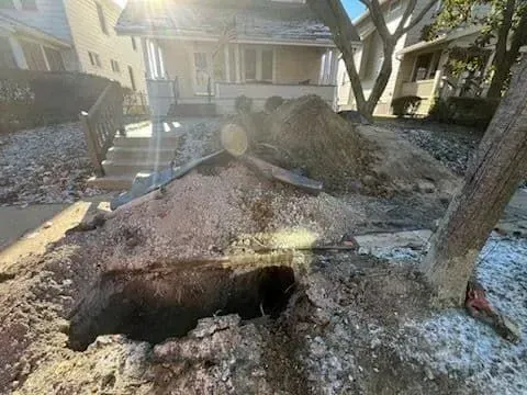 A deep trench dug in a lawn near a sidewalk, with dirt piles and wooden supports.