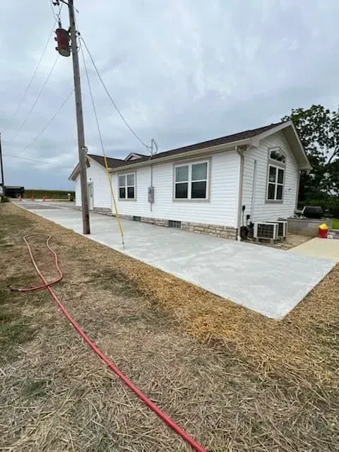 White house with new concrete patio, power lines, and red hose in a rural setting.