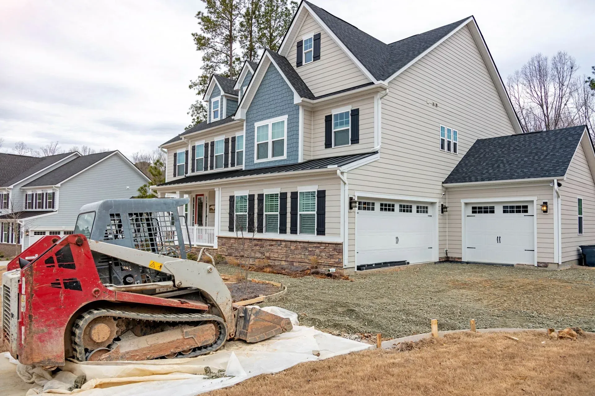 Two-story house under construction, beige siding with blue gable, red skid steer, gravel lot.