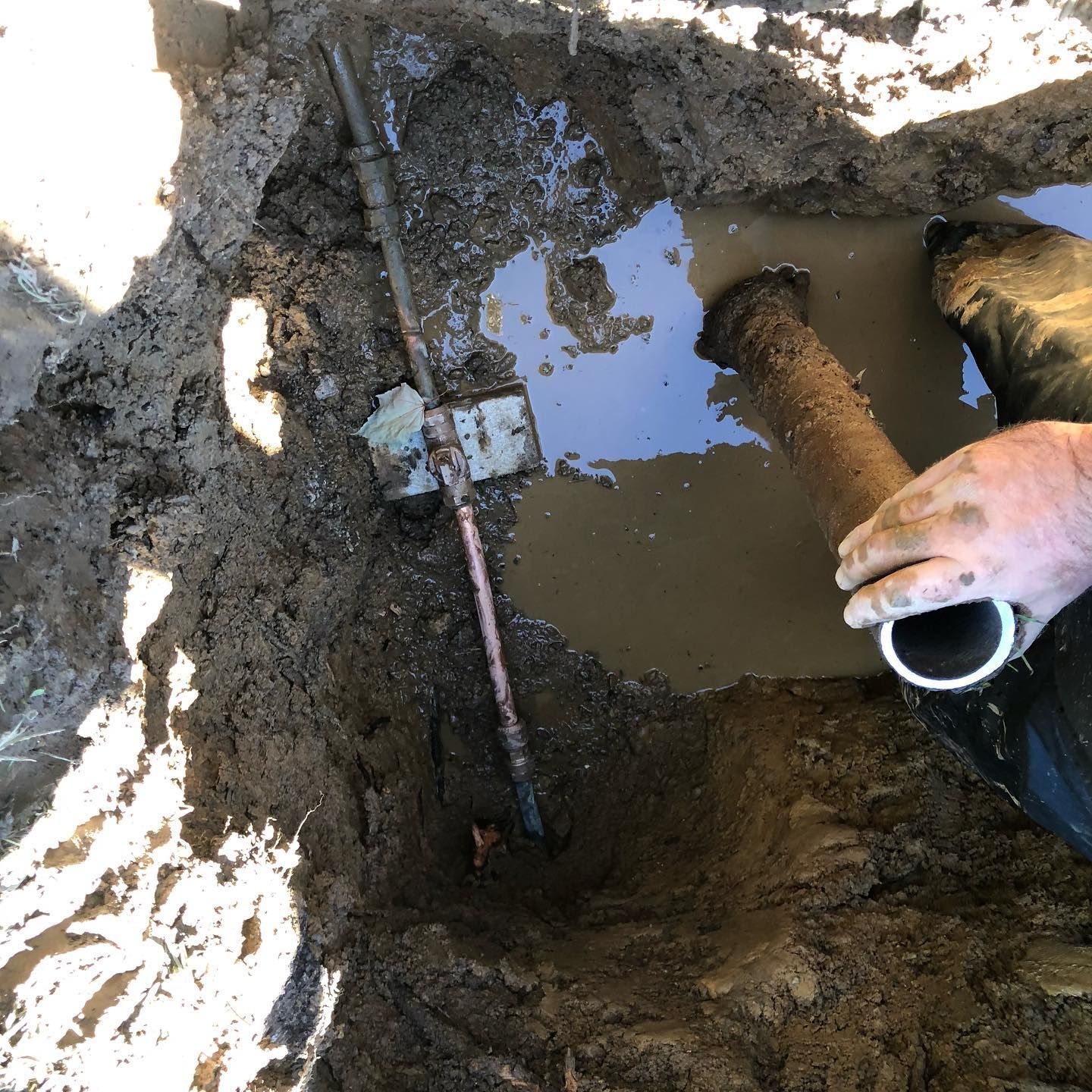 A hole in the ground filled with muddy water, exposing pipes. A person holds a pipe, hands covered in dirt.
