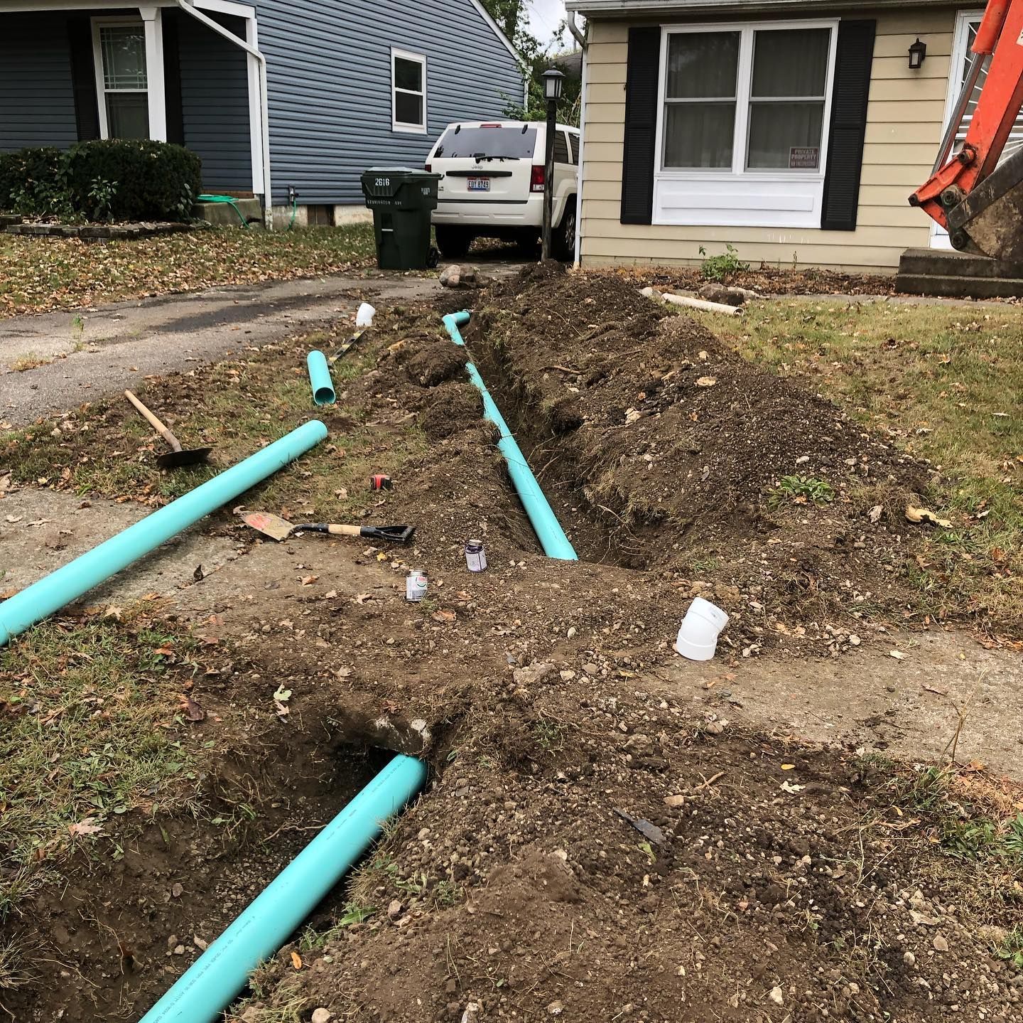 A construction site with blue pipes in trenches across a driveway. A backhoe and a house are visible.