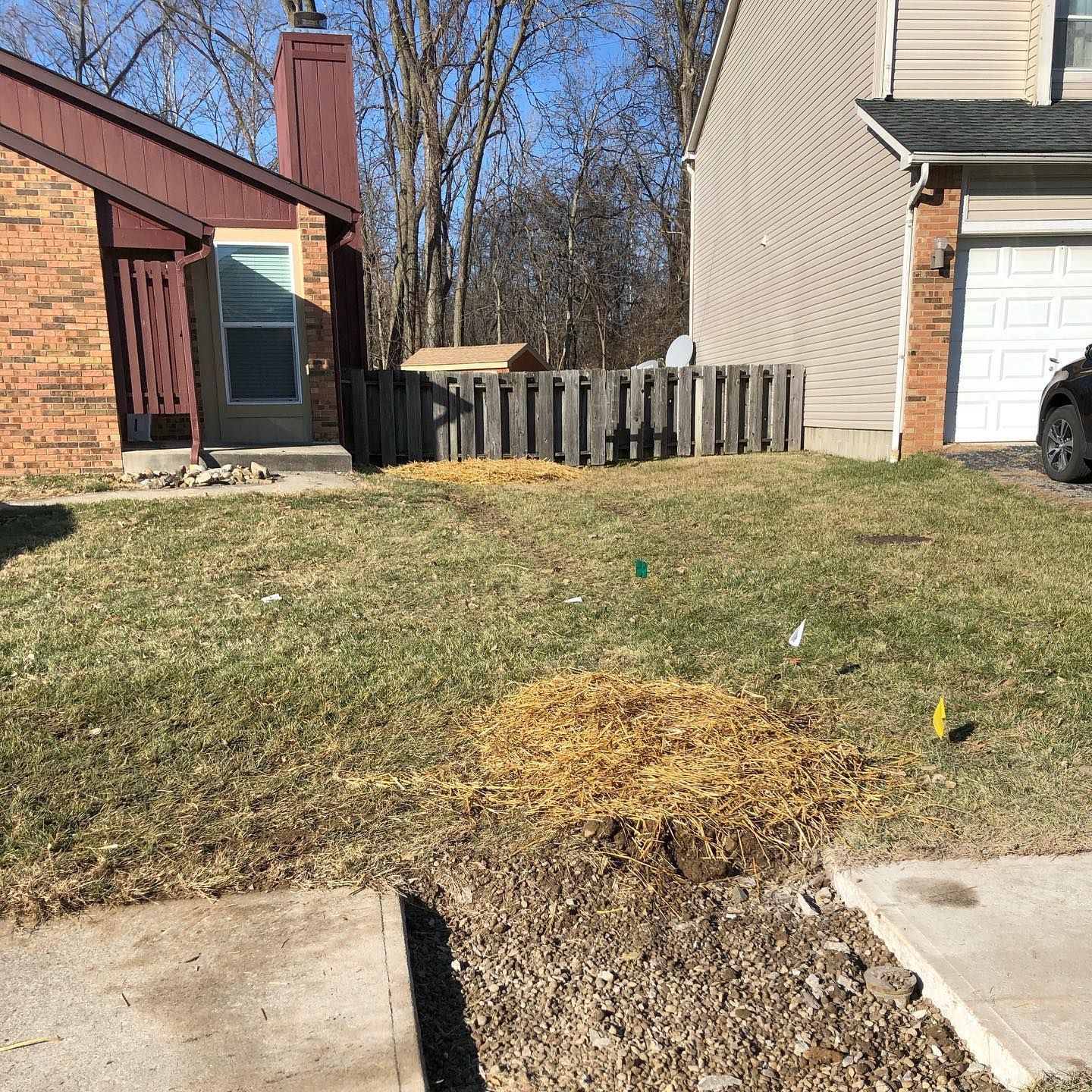 Two people in a trench, working near a buried pipe. One wears orange, one blue. Soil and shovel visible.
