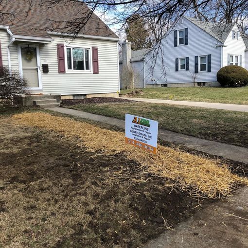 Man shovels dirt in a square pit, excavator in background, dirt pile, sunny day.