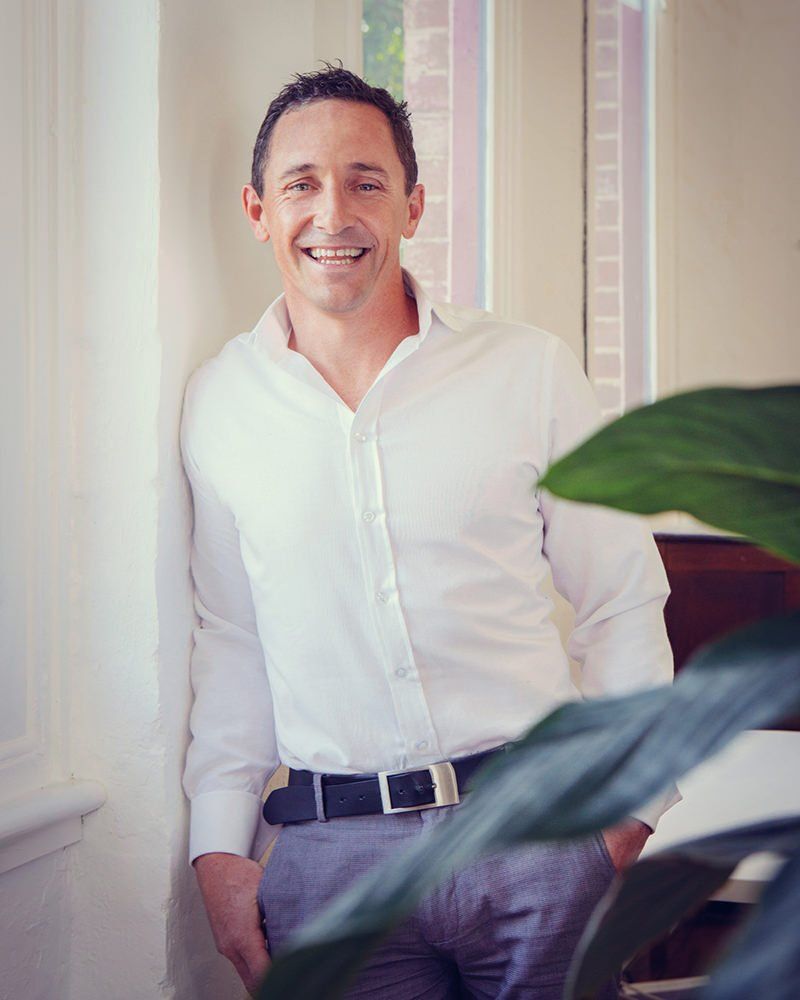 A man in a white shirt is leaning against a wall and smiling.