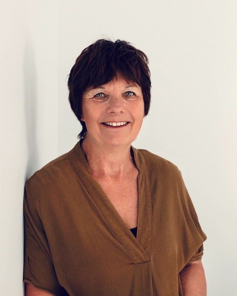 A woman in a brown shirt is leaning against a wall and smiling.