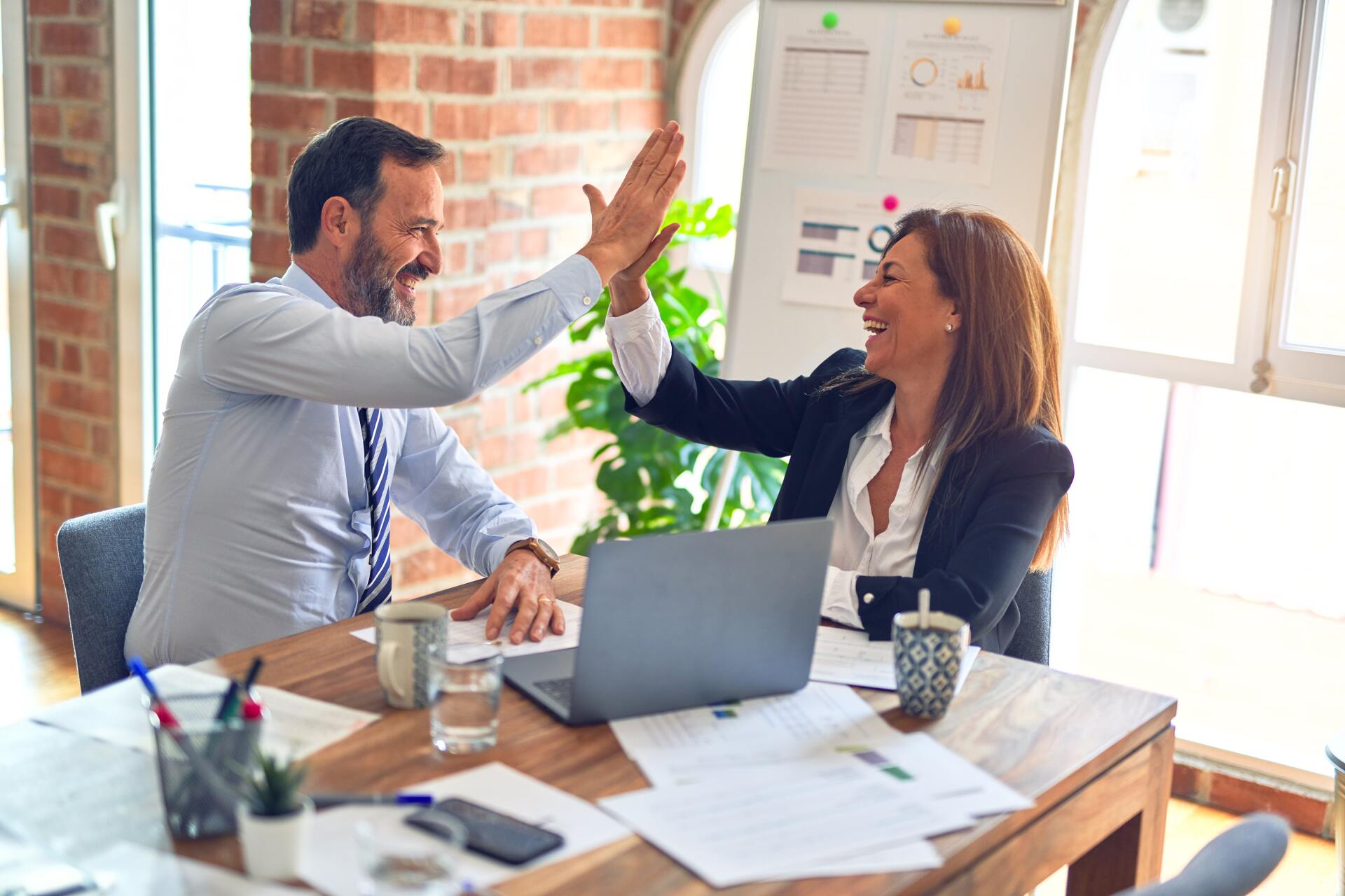 A man and a woman in an office high fiving each other at a desk with papers and a laptop on it.