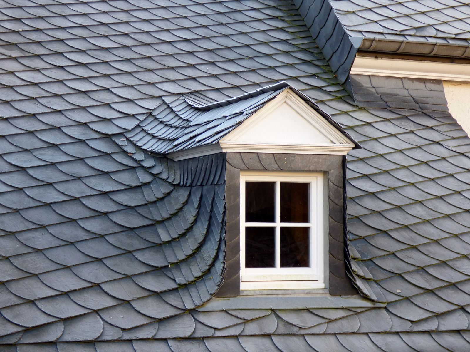 Dormer window with white trim on a slate roof, curved shingles around the window.