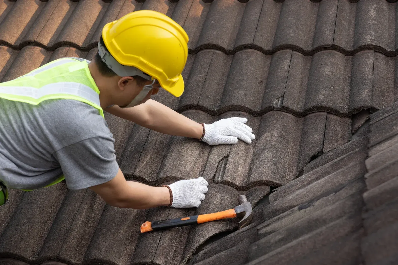 Worker in yellow hard hat repairing a shingled roof with a hammer.