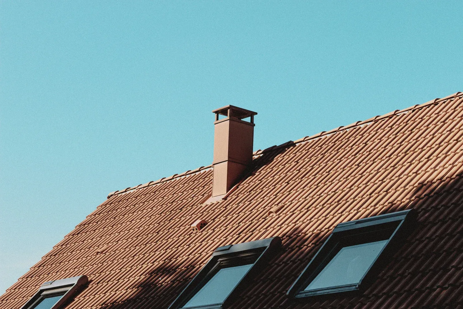 Red tiled roof with chimney and skylights against a clear blue sky.