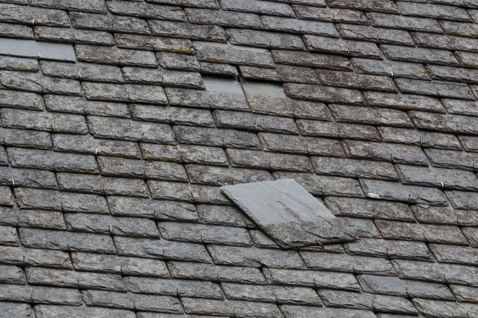 Damaged gray asphalt shingle roof, with several missing and loose tiles.