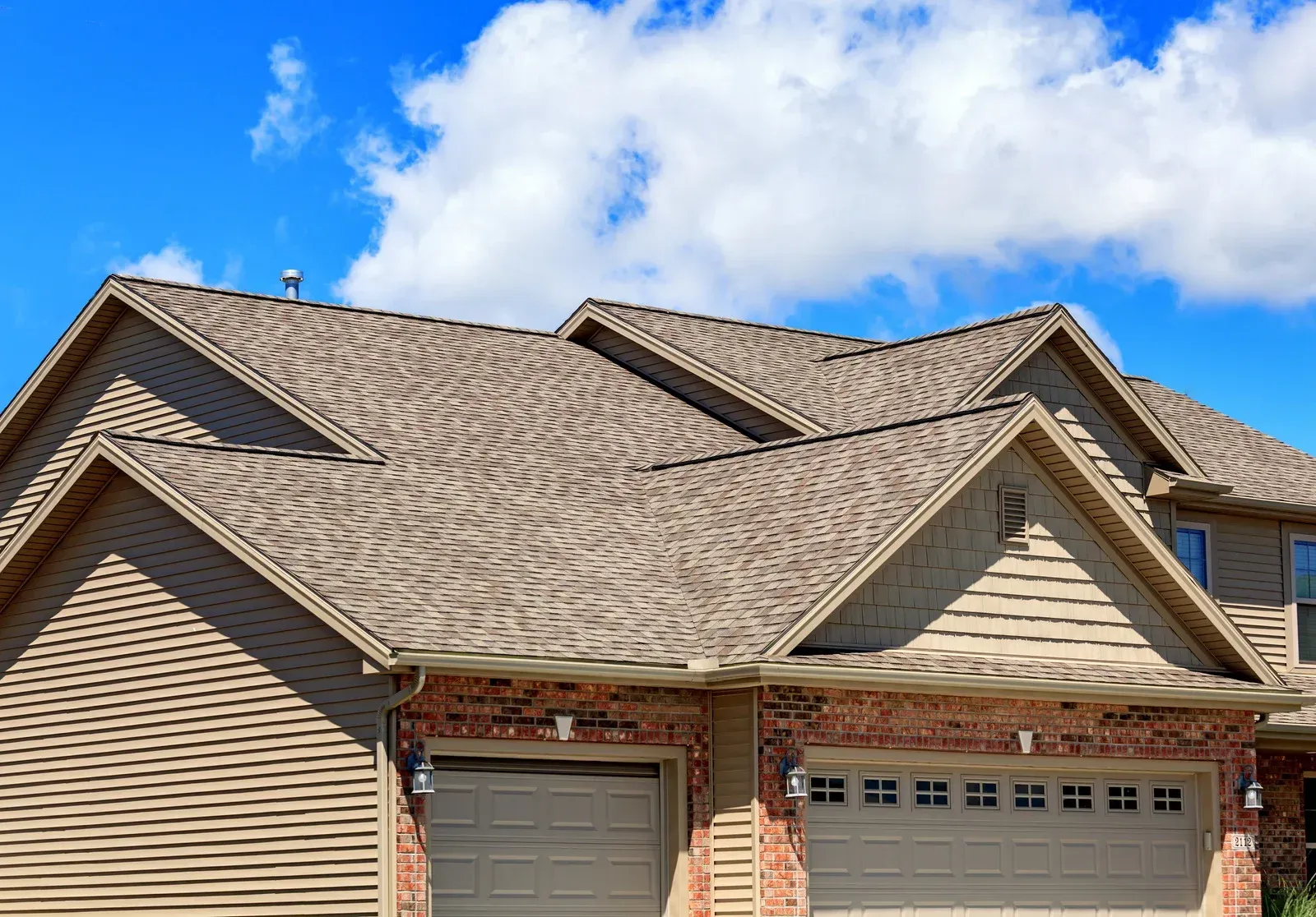 Suburban house with beige siding, tan shingle roof, and two garage doors under a blue sky