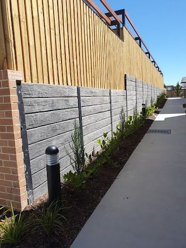 Stone patio leads to a bark-covered play area near a fence and building, under a blue sky.