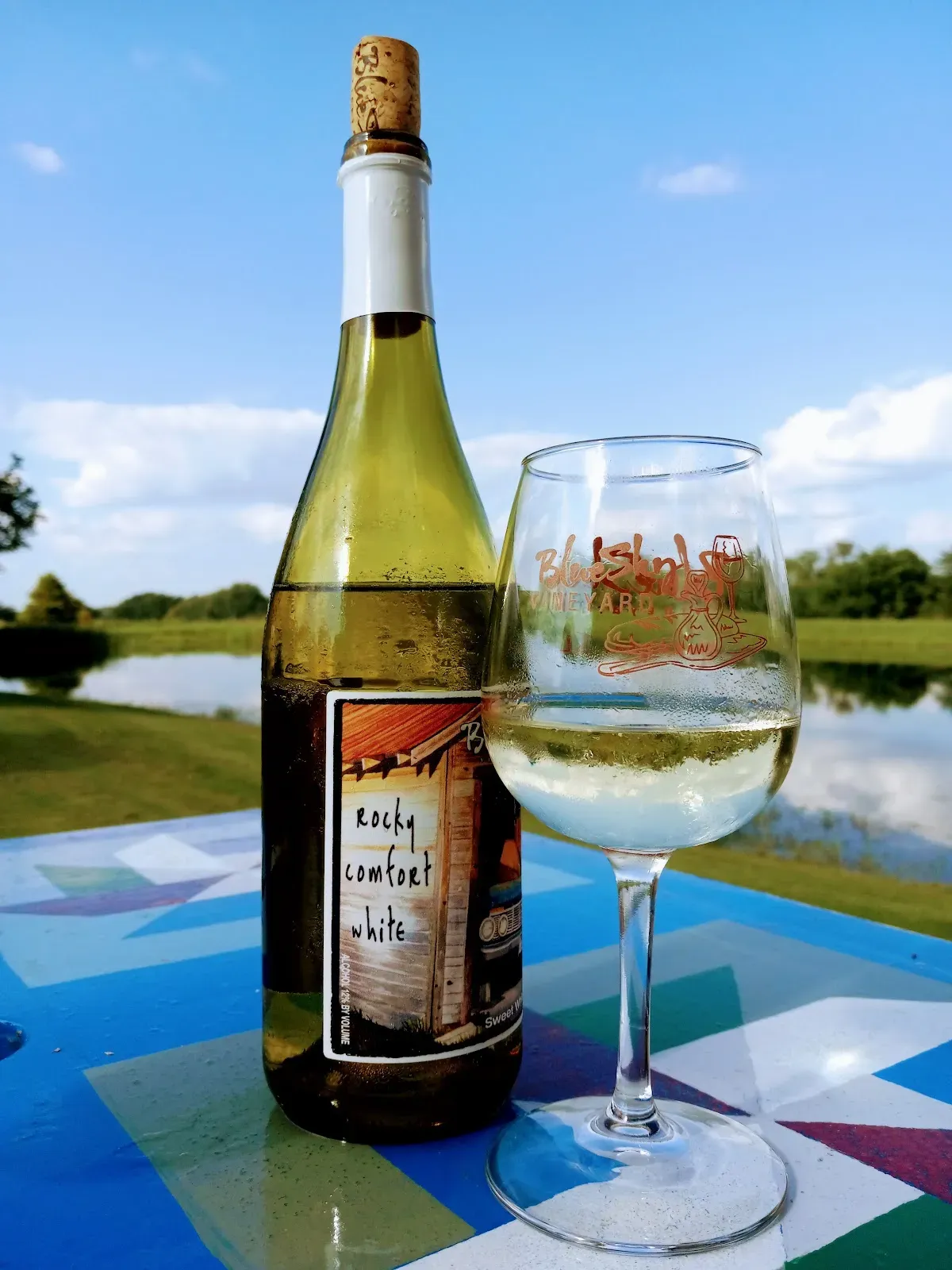 Bottle of white wine and a filled glass on a table outdoors, with a pond and trees in the background.