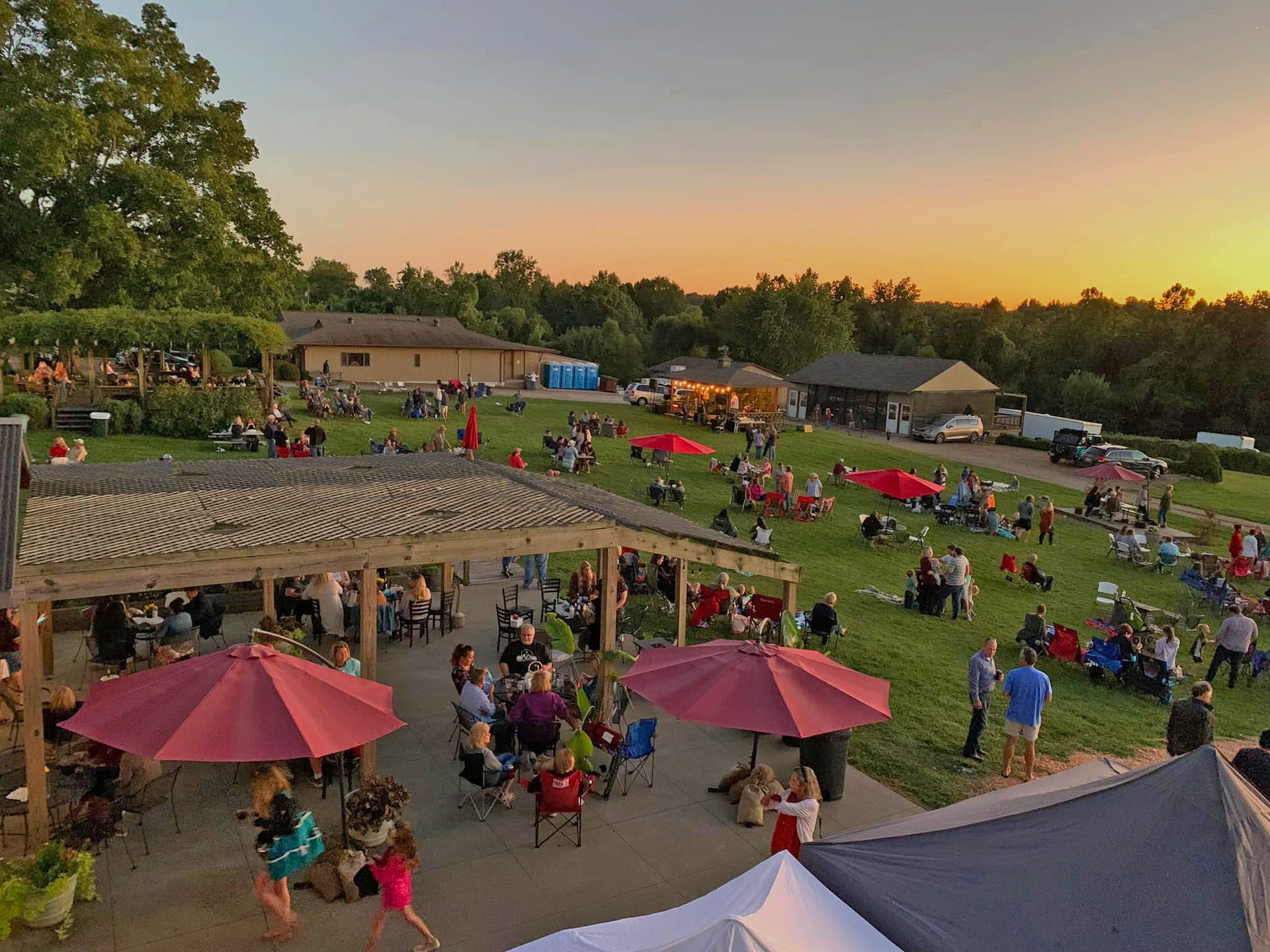 People enjoying an outdoor event with red umbrellas, tables, and a sunset backdrop.