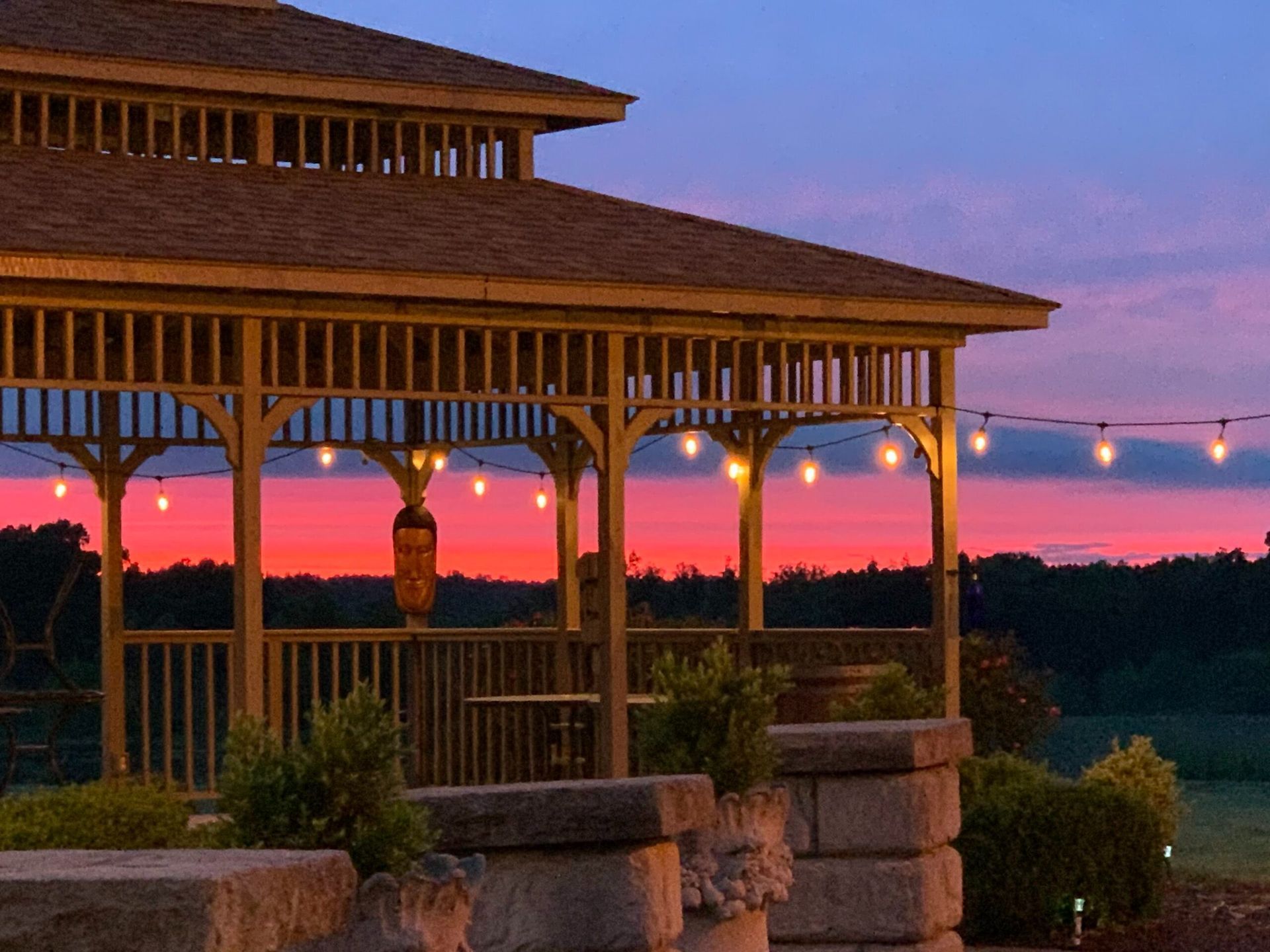 Gazebo at sunset with string lights. Pink and purple sky over trees.