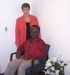 Woman in a red blazer stands behind a Black man seated in a chair, both smiling indoors. White flowers in the corner.