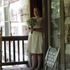 Woman in a white dress holding flowers stands on a porch. Rustic wood frame, folding chair nearby.