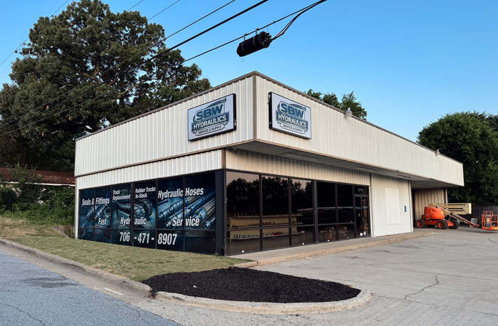 Corner auto shop with large windows, white siding, and blue signage beside a road.