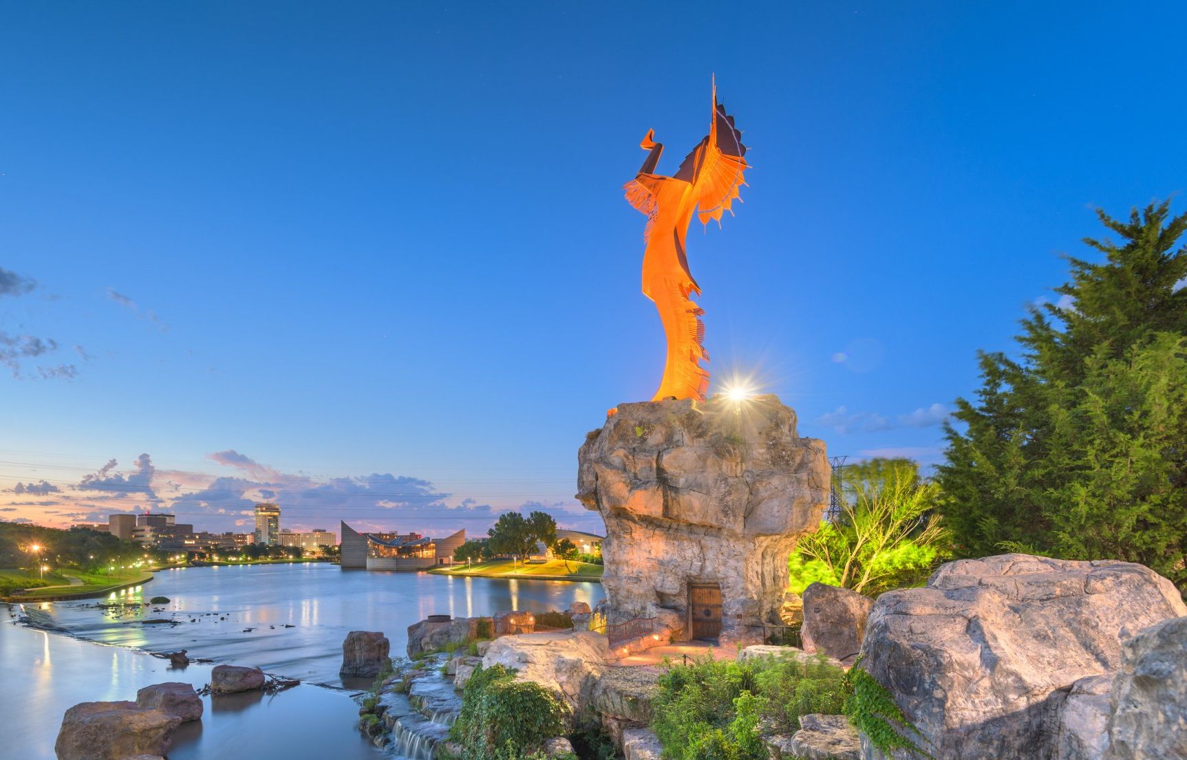 A statue of the keeper of the planes in Wichita, Kansas standing on top of a rock next to the Arkansas river.