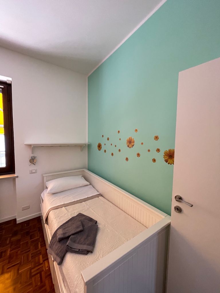 Piccola camera da letto con un letto singolo contro una parete verde acqua con adesivi floreali. Porta bianca sulla destra.