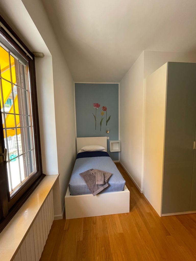 Piccola camera da letto con letto singolo, parete azzurra con fiori, pavimento in legno e ampia finestra.