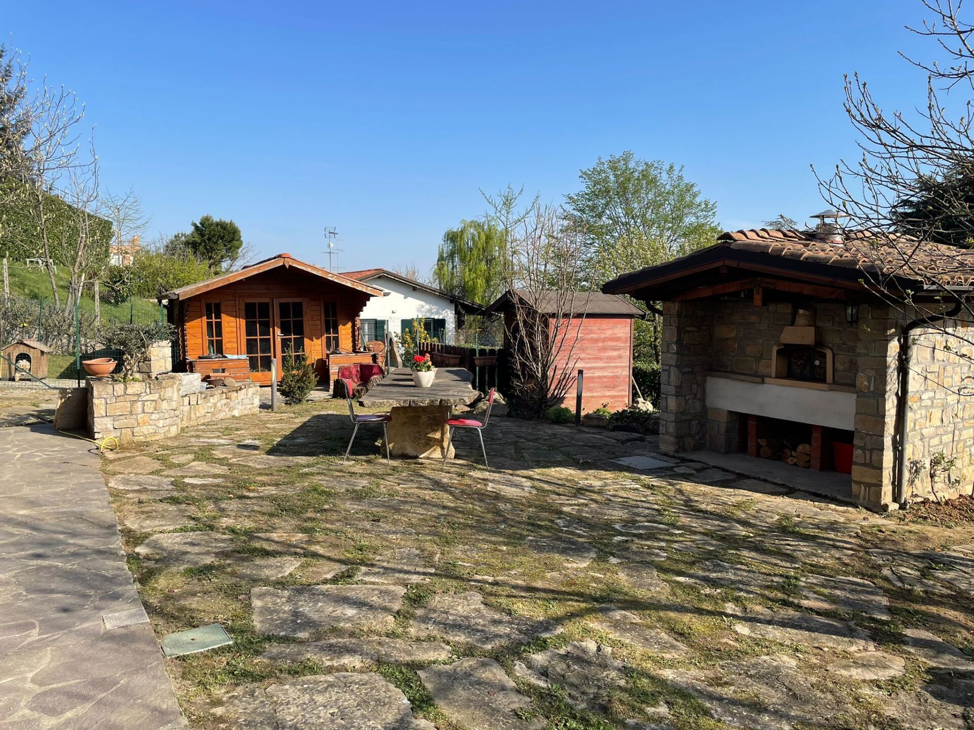 Un cortile soleggiato con una tettoia in legno, un forno in pietra e piccoli edifici sotto un cielo azzurro.