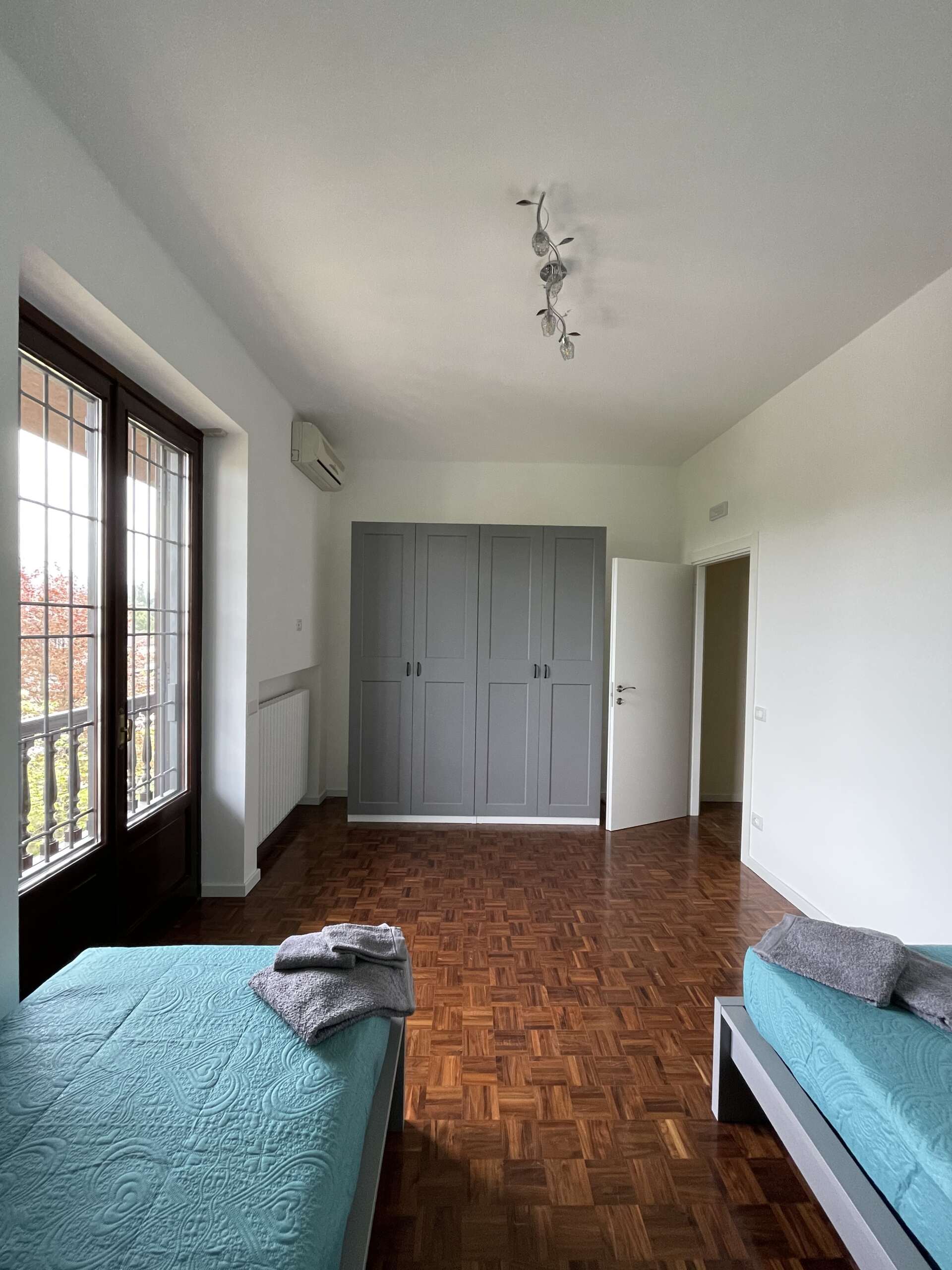 Camera da letto con due letti blu, armadio grigio, pavimento in parquet e finestra con balcone.