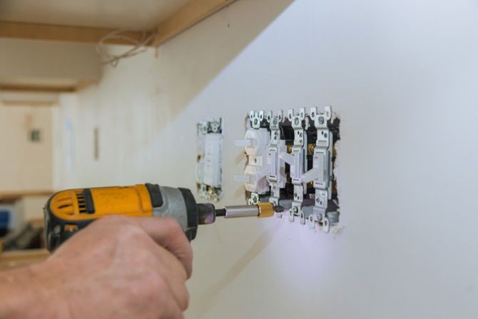 Hand drilling into a wall outlet box in a white hallway
