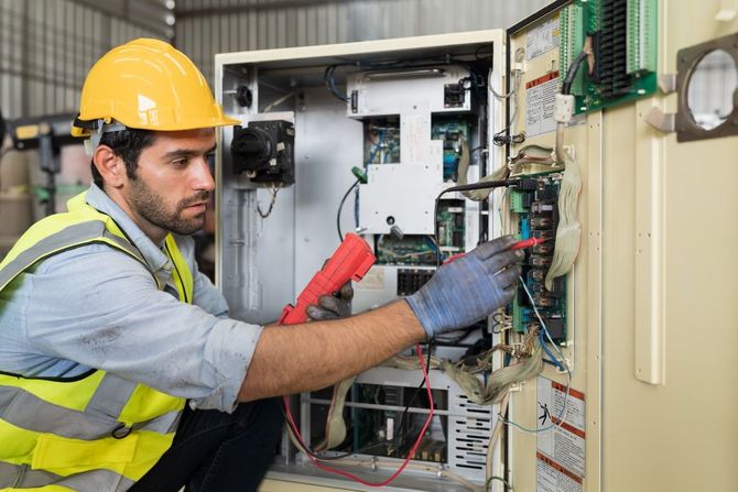 Technician in hard hat inspects electrical panel with tools inside an open control cabinet