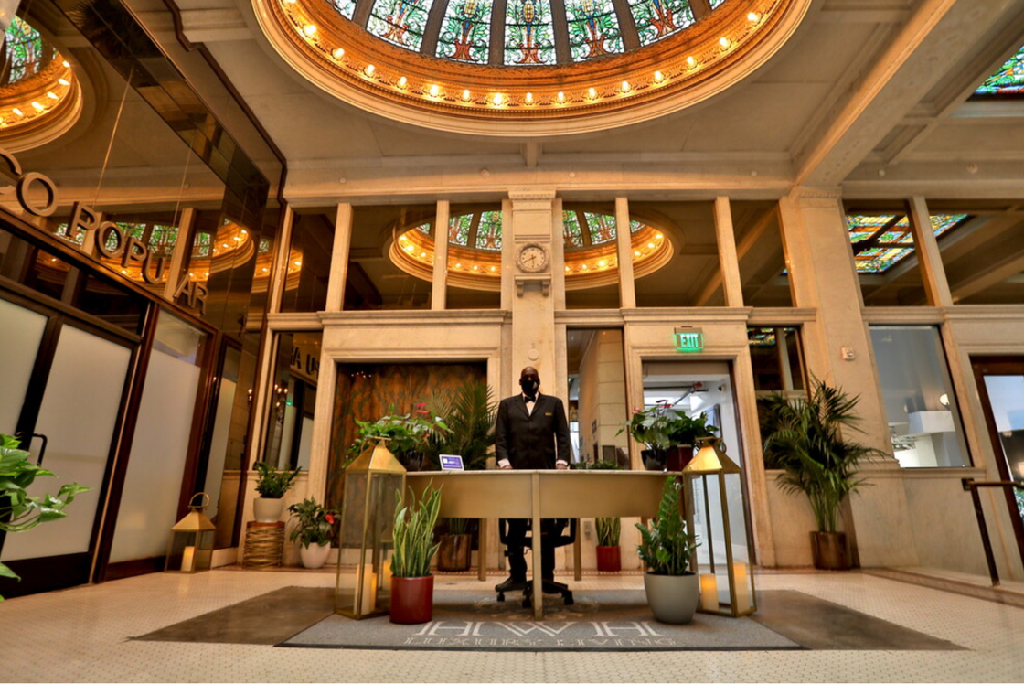 A grand, historic lobby at HWH Luxury Living in DTLA/Downtown Los Angeles featuring a 24-hour front desk with a concierge. The space showcases a stunning ornate stained-glass dome ceiling, golden architectural accents, and classical columns, blending historic HW Hellman Building charm with modern luxury.