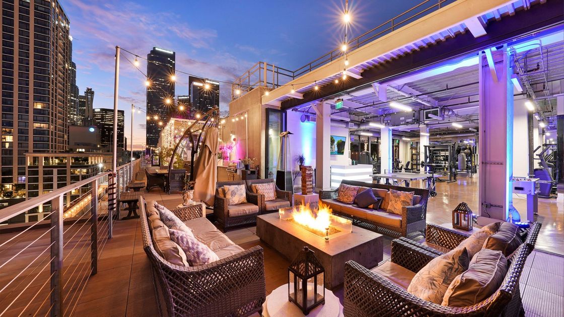 The expansive outdoor rooftop lounge at HWH Luxury Living in DTLA at dusk. The terrace features luxury wicker sectional seating, a modern stone fire pit, and string lights, set against a backdrop of glowing city skyscrapers and a sunset sky.
